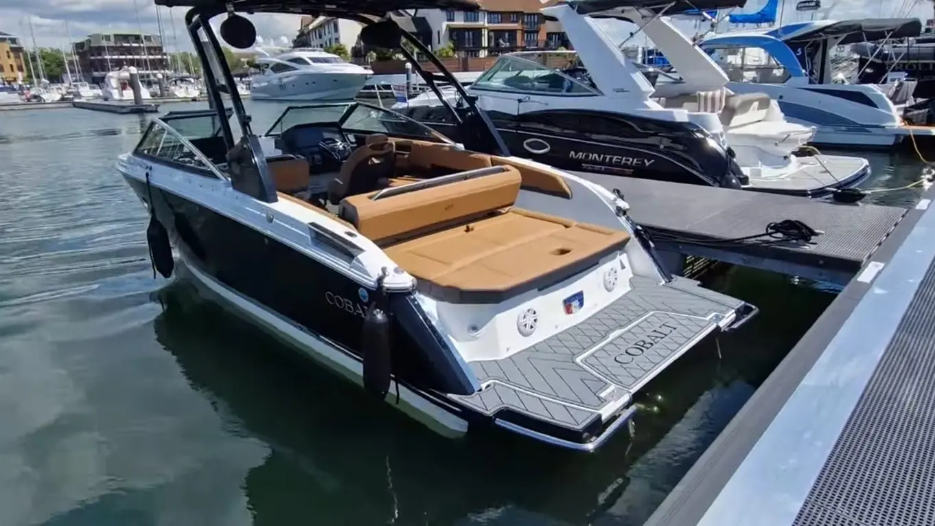 A Cobalt boat docked by a marina, featuring a tan seating area and nearby vessels, under a bright blue sky.